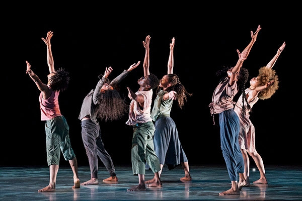 A group of 6 dancers facing the left with their arms stretched to the sky.